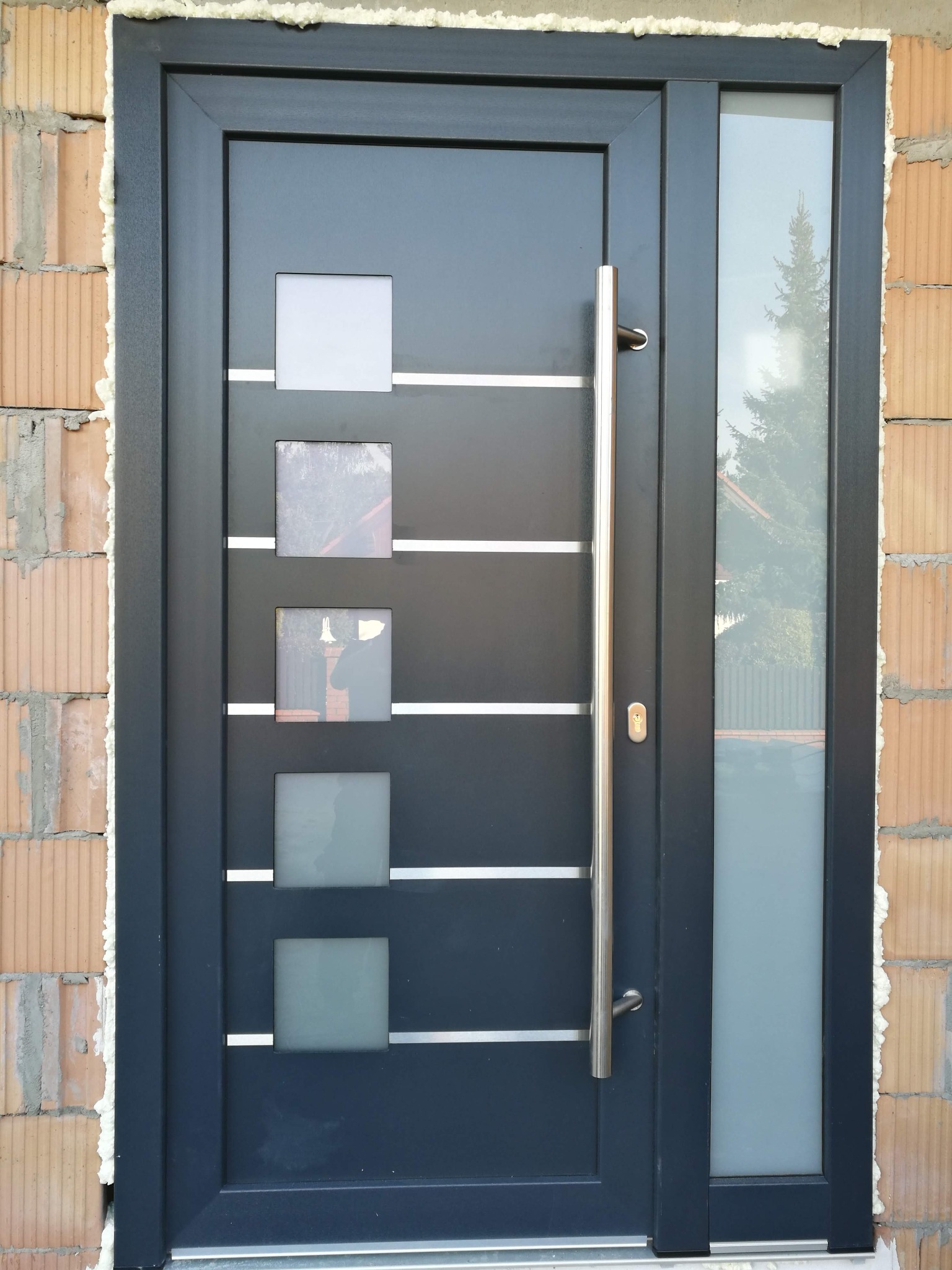 Nowoczesne, grafitowe drzwi wejściowe z czterema kwadratowymi przeszkleniami i długim, pionowym uchwytem, zamontowane w surowym murze z widoczną pianką montażową.