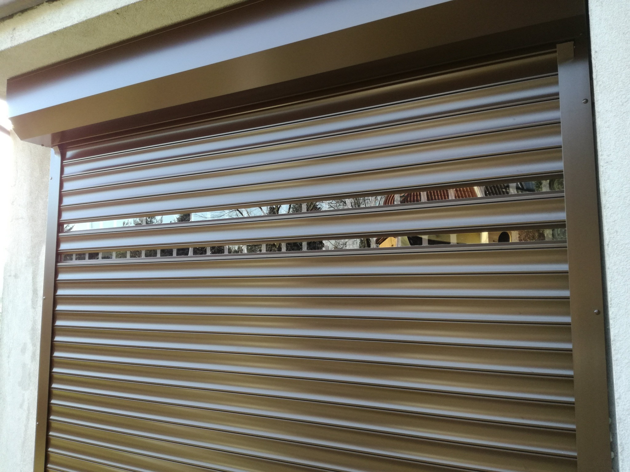 Częściowo opuszczona, brązowa roleta zewnętrzna z widocznymi poziomymi lamelami i odbiciem otoczenia w szczelinach.