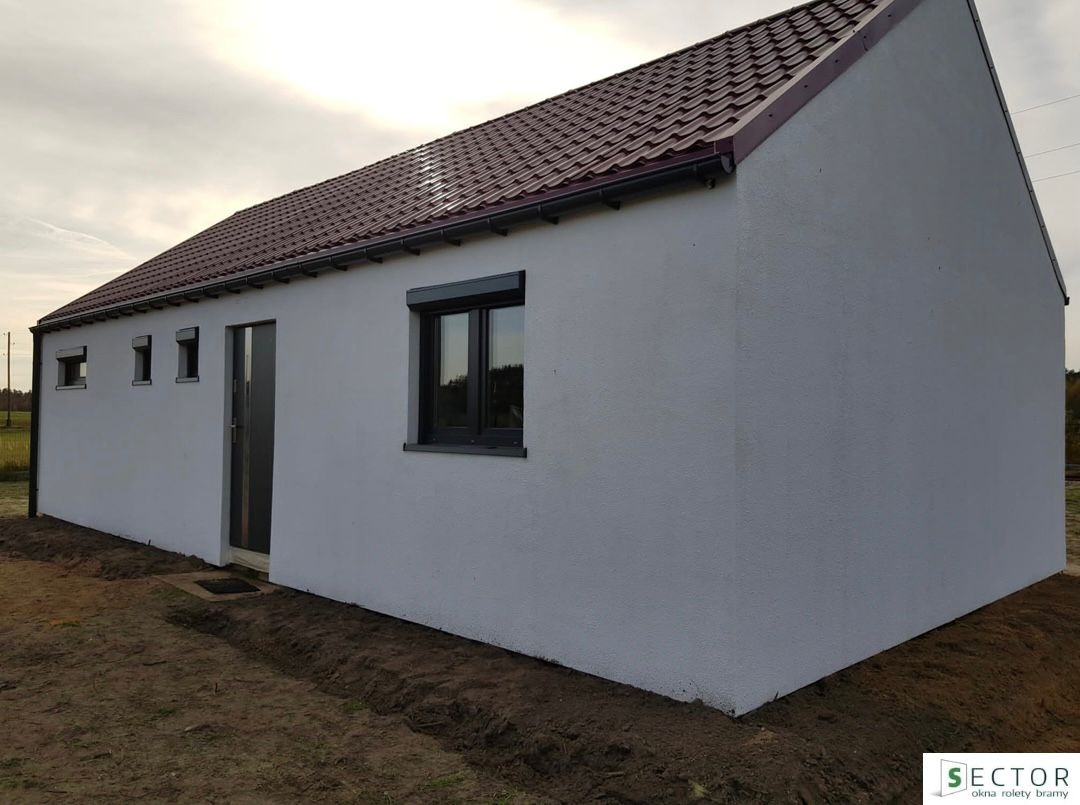 Nowoczesny budynek gospodarczy z szarą stolarką okienną i drzwiami, widoczne rolety zewnętrzne w oknach, świeżo obsypane ziemią obrzeża budynku, brązowa dachówka.