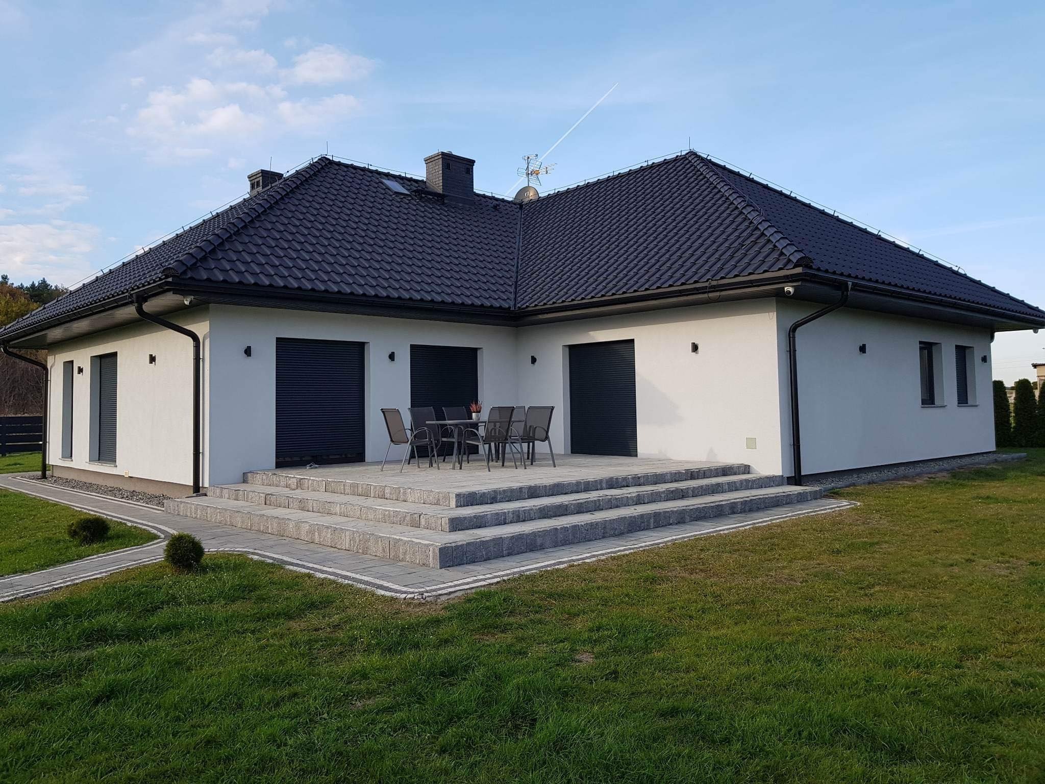 Nowoczesny dom jednorodzinny z ciemną dachówką, szarymi roletami zewnętrznymi, kamiennymi schodami prowadzącymi na taras z meblami ogrodowymi i zadbanym trawnikiem.