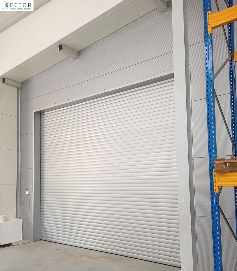 Zamknięta, biała, stalowa brama rolowana w hali magazynowej, widoczne regały z paletami w tle.