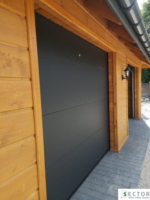 Ciemnoszara segmentowa brama garażowa z automatycznym zamkiem, zamontowana w drewnianym garażu z oświetleniem zewnętrznym.