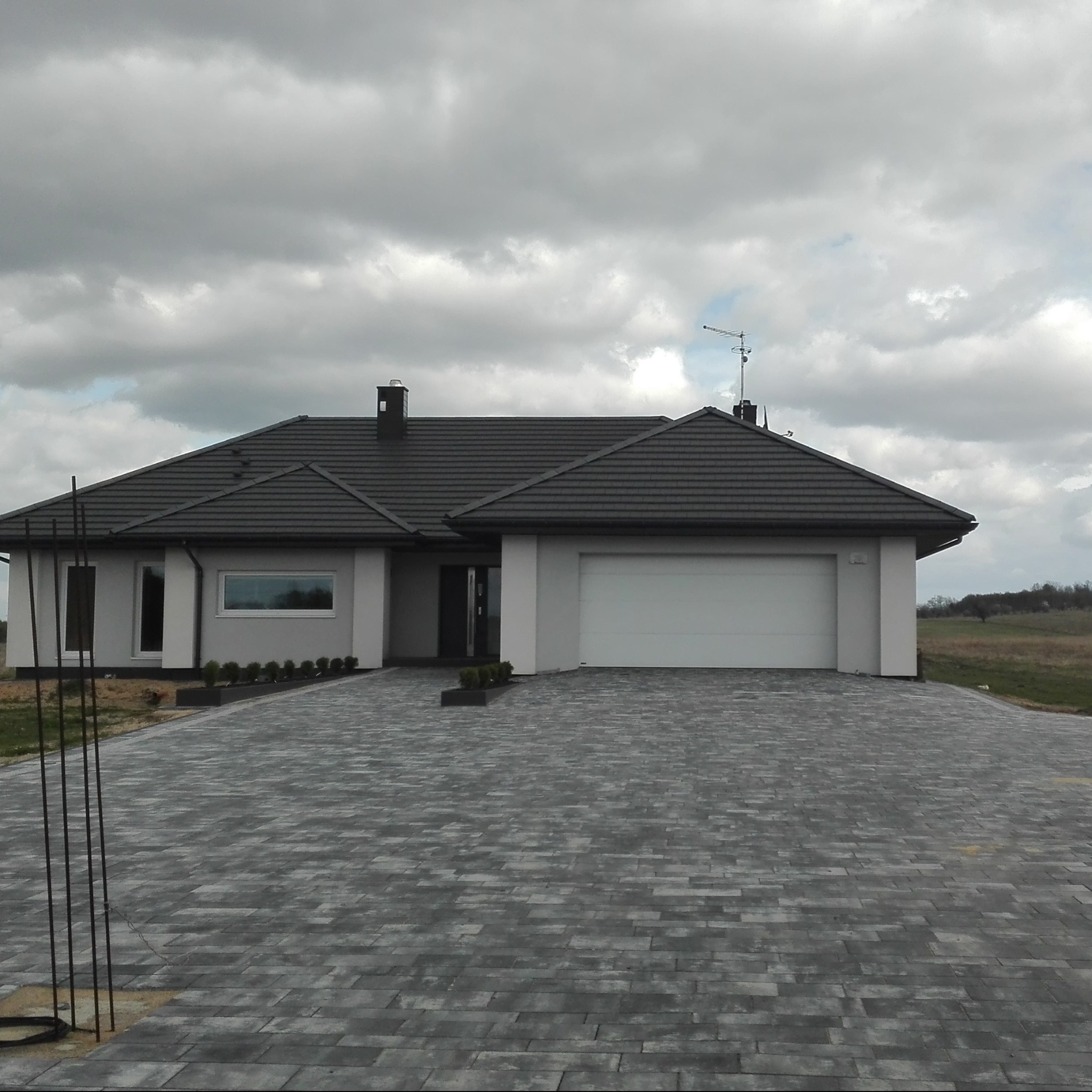 Nowoczesny dom jednorodzinny z szarą kostką brukową na podjeździe i białymi ścianami, widoczna brama garażowa i elementy instalacji antenowej na dachu.