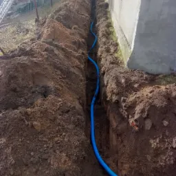 Wykopany wąski rów w ziemi, w którym ułożono niebieski karbowany przewód instalacyjny, widoczny fragment budynku i pracownicy w tle.