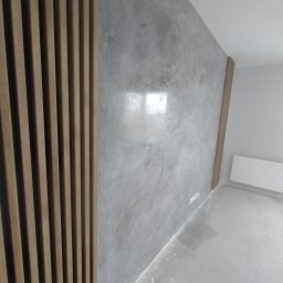 BOBI MAKER USŁUGI BUDOWLANE - Ściana z szarą, dekoracyjną fakturą, częściowo wykończona drewnianymi lamelami, widoczny grzejnik i gniazdka elektryczne.