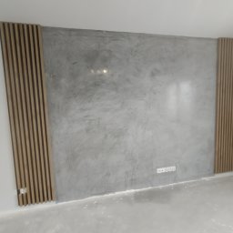 BOBI MAKER USŁUGI BUDOWLANE - Ściana z szarą, dekoracyjną fakturą tynku w stylu betonu architektonicznego, ozdobiona pionowymi panelami lamelowymi po bokach, widoczne gniazdka elektryczne.