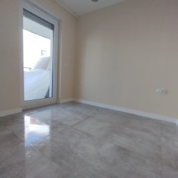 BOBI MAKER USŁUGI BUDOWLANE - Wykończone wnętrze pokoju z szarymi płytkami podłogowymi, widok na balkon przez otwarte drzwi balkonowe, białe listwy przypodłogowe i lampa sufitowa w geometrycznym stylu.