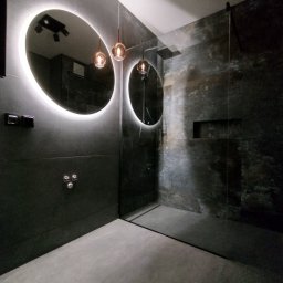 BOBI MAKER USŁUGI BUDOWLANE - Nowoczesna łazienka z ciemnymi płytkami, okrągłymi lustrami z podświetleniem LED i szklaną kabiną prysznicową typu walk-in, oświetlona wiszącymi lampami z miedzianymi detalami.