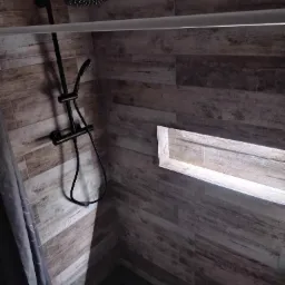 Nowoczesna kabina prysznicowa z czarną armaturą, wykończona płytkami imitującymi drewno i podświetlaną wnęką w ścianie.