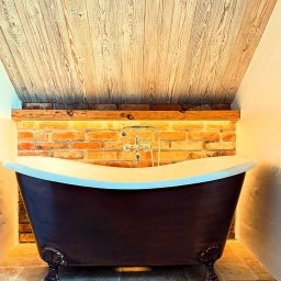 BOBI MAKER USŁUGI BUDOWLANE - Stylowa łazienka z wolnostojącą wanną na ozdobnych nogach, ceglaną ścianą z podświetlaną drewnianą półką i skośnym sufitem z bielonego drewna.