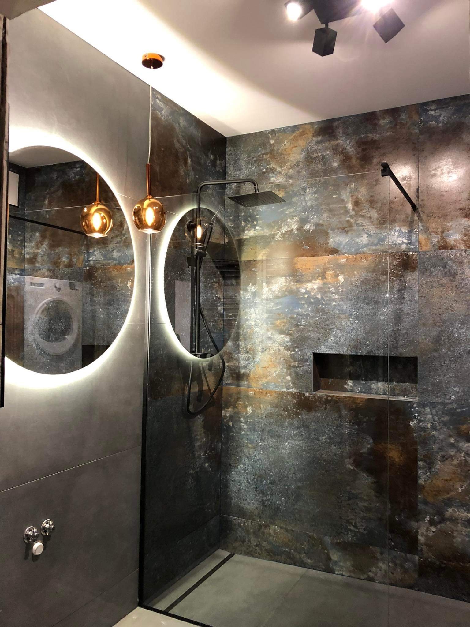 Nowoczesna łazienka z prysznicem typu walk-in, wykończona płytkami imitującymi rdzę, oświetlona okrągłymi lustrami z podświetleniem LED i wiszącymi lampami w kształcie kul, z widoczną pralką...