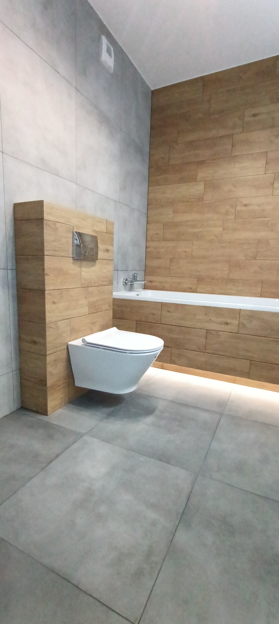 Nowoczesna łazienka z podwieszaną toaletą, drewnopodobne płytki na ścianach i podświetleniem LED pod wanną, szare płytki podłogowe.