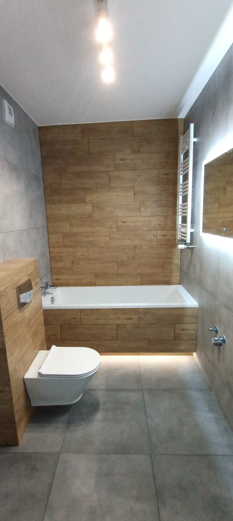 Nowoczesna łazienka z drewnopodobnymi płytkami na ścianie, podświetlana wanna i podwieszana toaleta, szare płytki podłogowe, minimalistyczny grzejnik drabinkowy i lustro z podświetleniem LED.