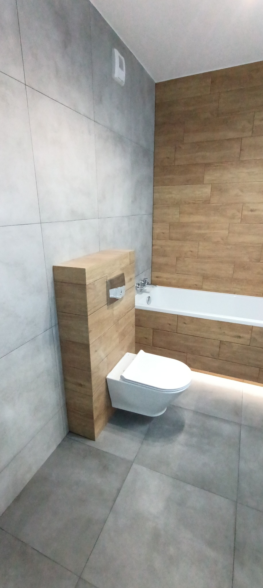 Nowoczesna łazienka z podwieszaną toaletą, obudowaną drewnopodobną płytką, szare płytki na ścianach i podłodze, wanna z drewnopodobną obudową i podświetleniem LED.