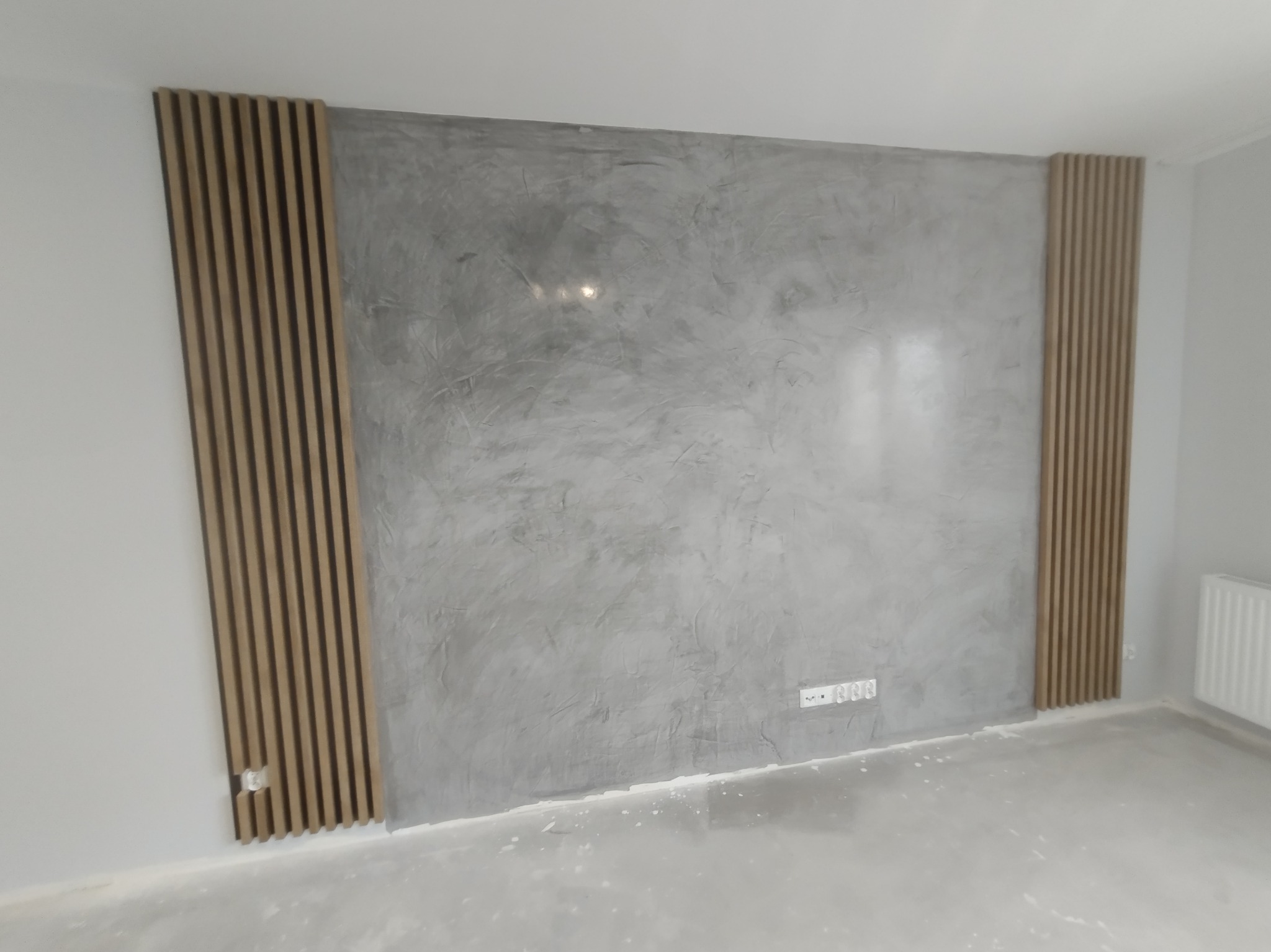 Ściana z szarą, dekoracyjną fakturą tynku w stylu betonu architektonicznego, ozdobiona pionowymi panelami lamelowymi po bokach, widoczne gniazdka elektryczne.
