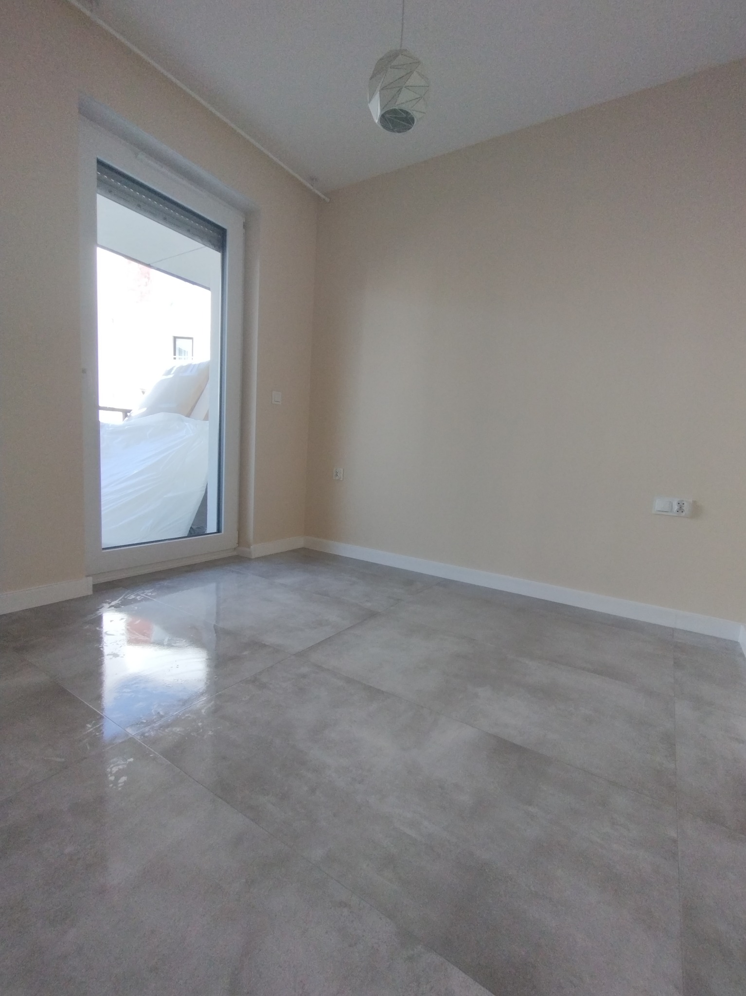 Wykończone wnętrze pokoju z szarymi płytkami podłogowymi, widok na balkon przez otwarte drzwi balkonowe, białe listwy przypodłogowe i lampa sufitowa w geometrycznym stylu.