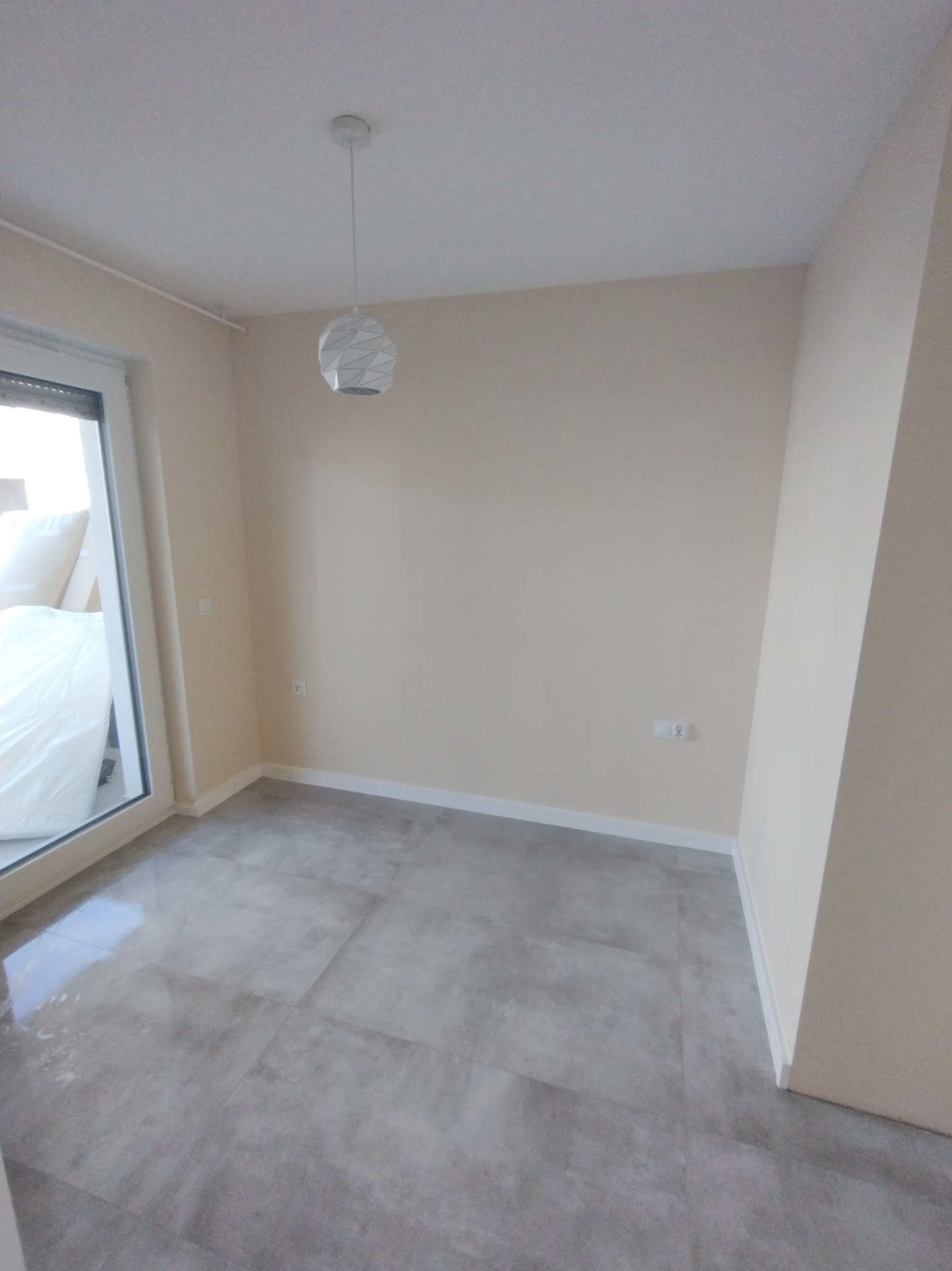 Pusty pokój po remoncie z szarymi płytkami podłogowymi, świeżo pomalowanymi ścianami w odcieniach beżu i nowoczesną, geometryczną lampą wiszącą.
