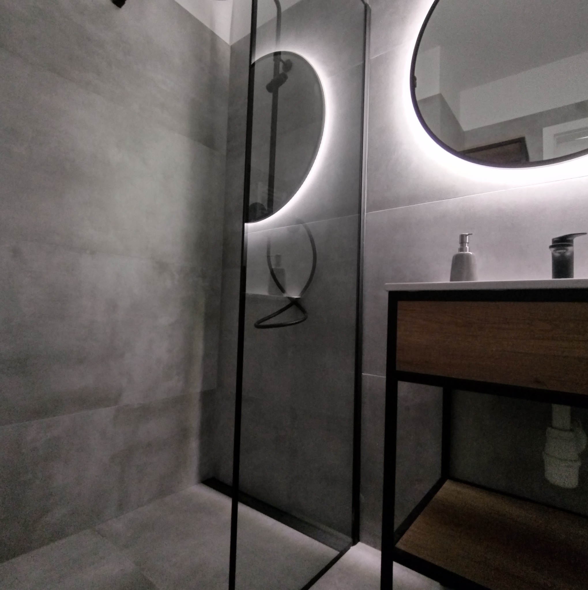 Nowoczesna łazienka w Malborku z szarymi płytkami, kabiną prysznicową ze szklaną ścianą, okrągłym lustrem z podświetleniem LED i umywalką z drewnianą szafką.