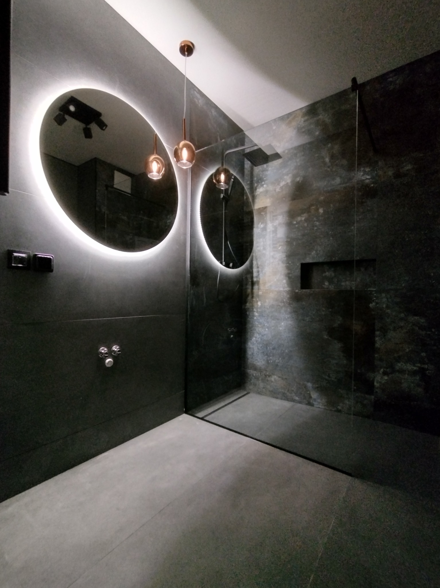Nowoczesna łazienka z ciemnymi płytkami, okrągłymi lustrami z podświetleniem LED i szklaną kabiną prysznicową typu walk-in, oświetlona wiszącymi lampami z miedzianymi detalami.
