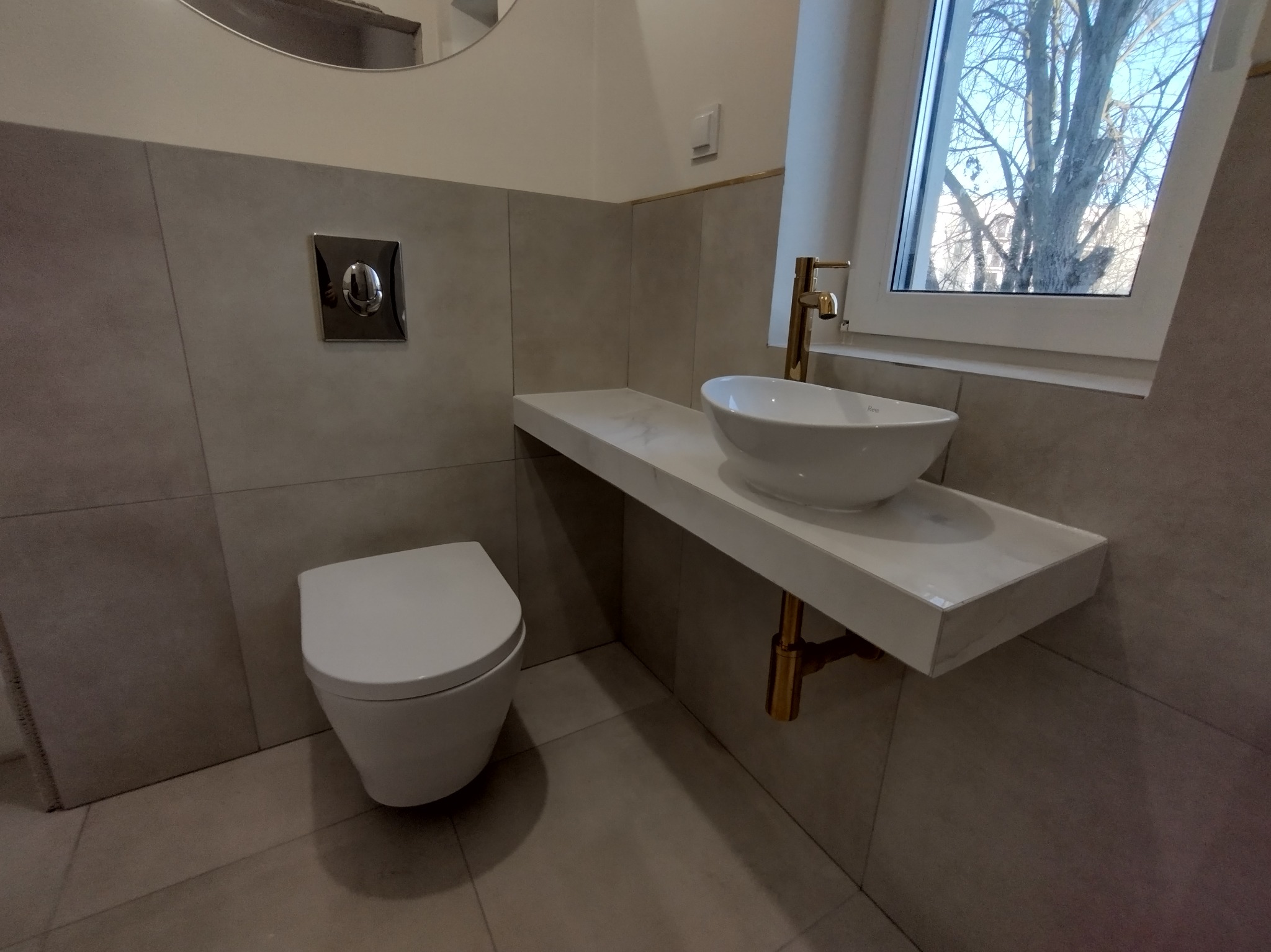 Nowoczesna łazienka z podwieszaną miską WC, umywalką nablatową na białym blacie i złotą armaturą, widok przez okno na drzewo bez liści.