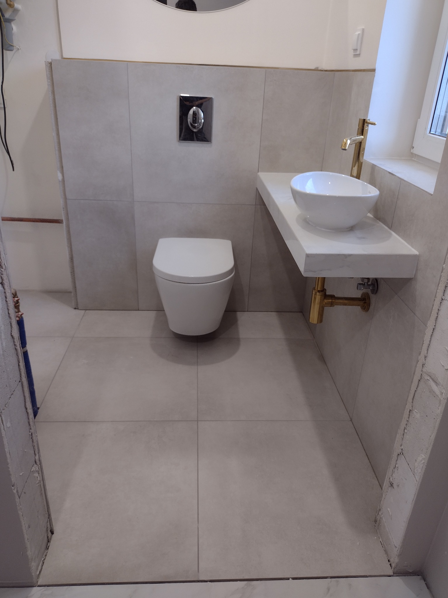Nowoczesna łazienka w trakcie wykańczania: podwieszana toaleta, umywalka nablatowa z baterią w kolorze złotym, szare płytki ścienne i podłogowe, widoczne niedokończone obrzeża przy drzwiach.