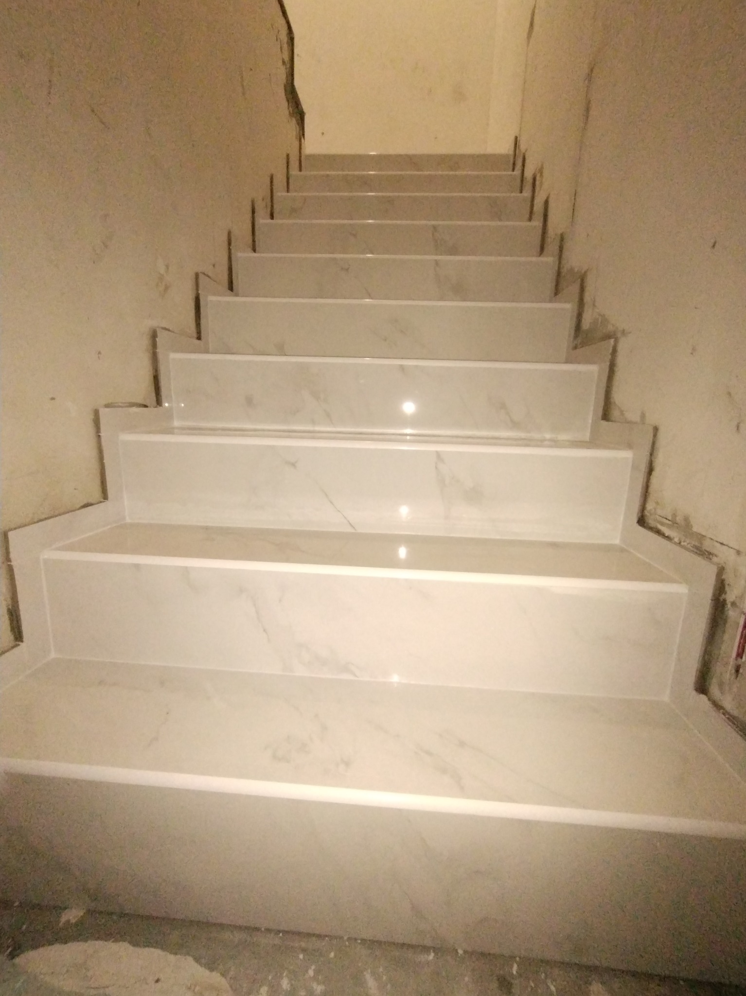 Schody wyłożone białymi płytkami imitującymi marmur, widoczne niedoskonałości na ścianach i podłodze wskazujące na trwający proces wykończeniowy.