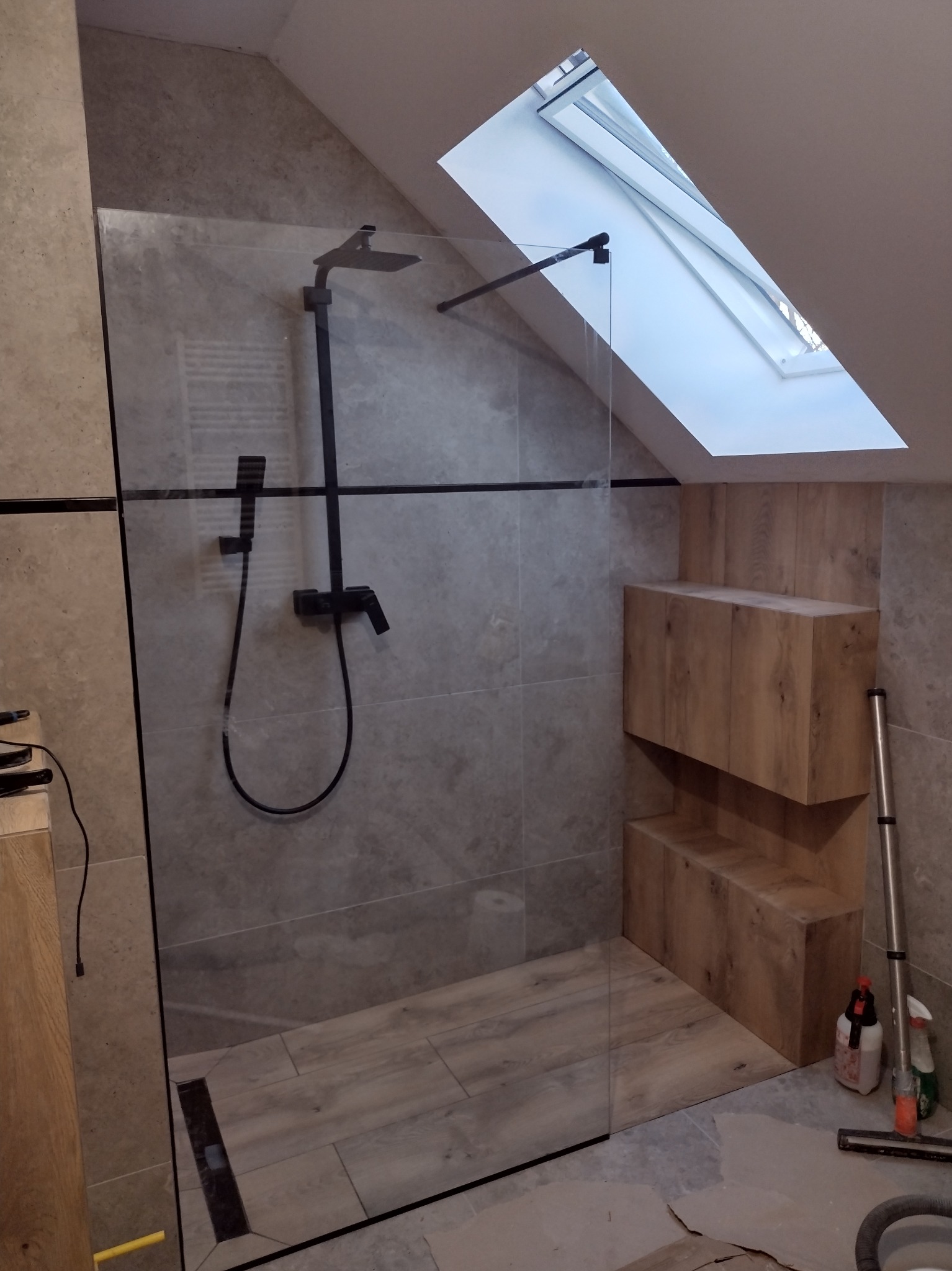 Nowoczesna kabina prysznicowa ze szklaną ścianą w łazience na poddaszu z oknem dachowym, czarną armaturą i drewnianymi półkami w zabudowie, widoczny odpływ liniowy w podłodze.