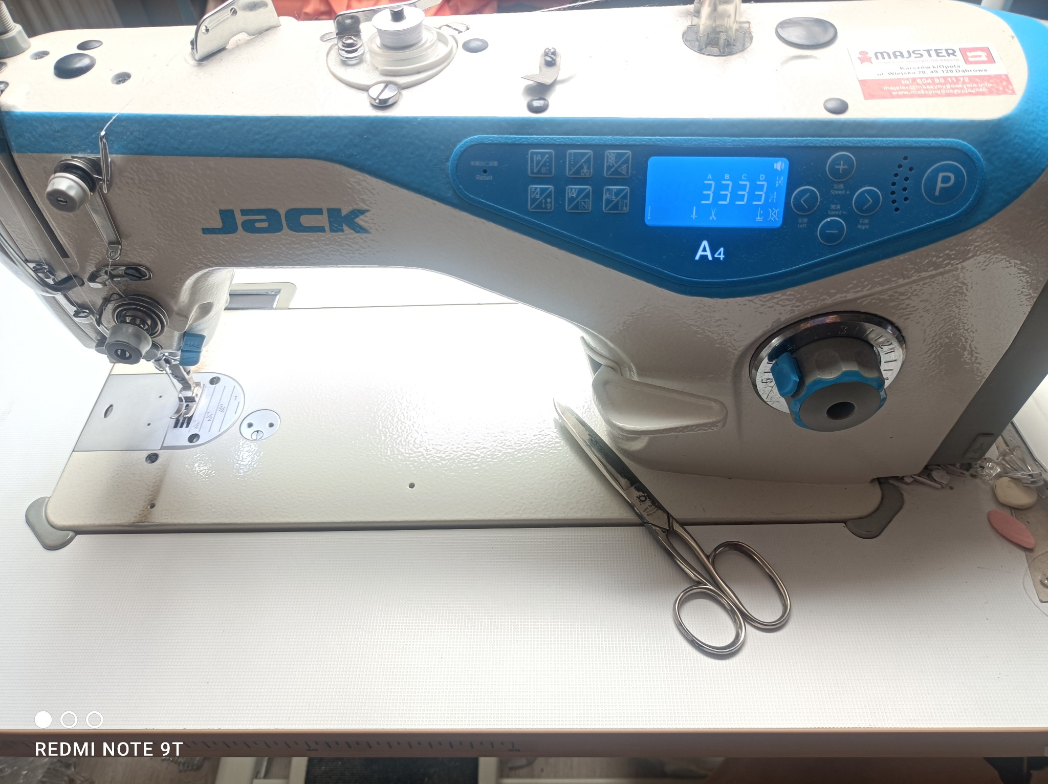 Przemysłowa maszyna do szycia marki Jack A4 z wyświetlaczem cyfrowym, nożyczkami obok i naklejką informacyjną Majster na obudowie.
