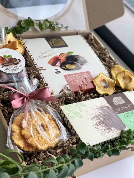 Zawartość kartonowego pudełka z ozdobnymi wypełniaczami: czekolady marki Laro, suszone ananasy w celofanie z różową wstążką, suszone jabłka i orzechy laskowe w czekoladzie.