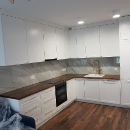 Nowoczesna kuchnia z białymi szafkami, drewnianym blatem i złotymi uchwytami, widok na zabudowę kuchenną z piekarnikiem i zlewozmywakiem, częściowo zasłonięta folią ochronną.