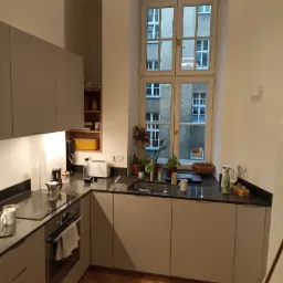 Kuchenne wnętrze z szarymi szafkami, ciemnym blatem, drewnianą podłogą w jodełkę i dużym oknem z widokiem na budynek z cegły