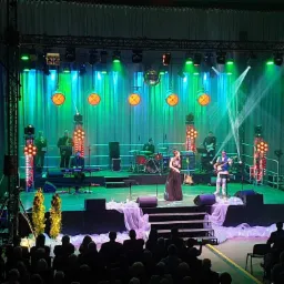 Zespół muzyczny na scenie podczas koncertu, oświetlenie sceniczne w kolorach zieleni i pomarańczy, publiczność siedząca na widowni.