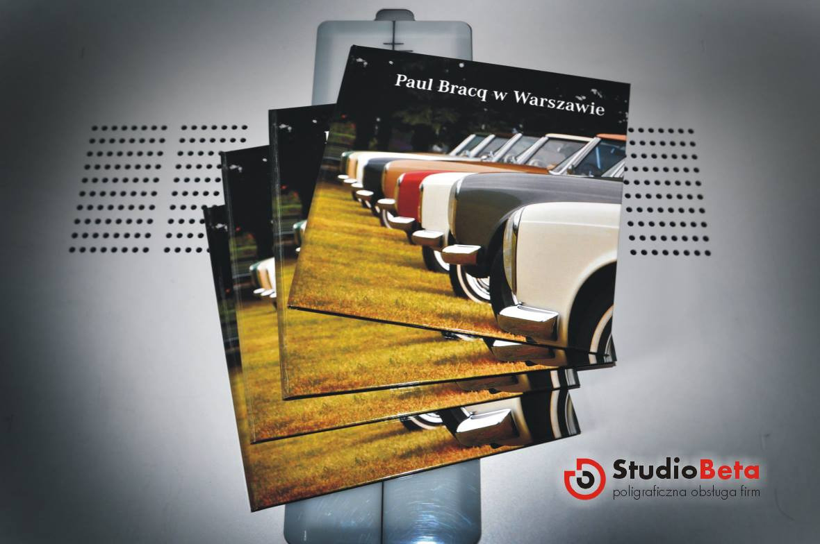 Stos wydrukowanych katalogów z okładką przedstawiającą rząd klasycznych samochodów na trawiastym tle, widoczne logo Studio Beta.