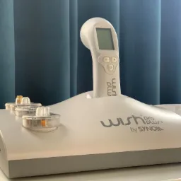 Urządzenie Wish Pro Plus by Synoia z aplikatorem i wymiennymi kapsułkami na białym stoliku, na tle zasłony w odcieniach niebieskiego.