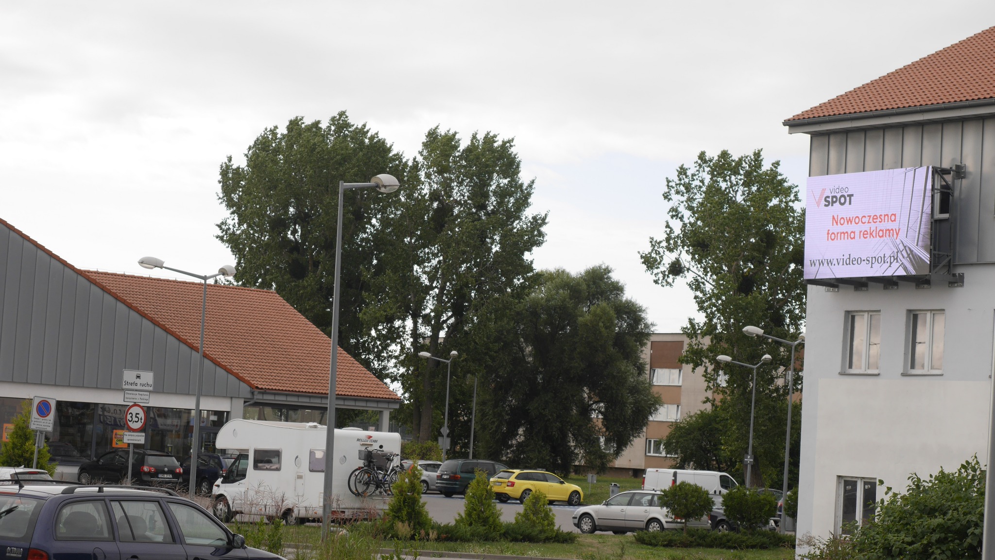 Zewnętrzny ekran LED z wyświetlaną reklamą na ścianie budynku, widok z ulicy z zaparkowanymi samochodami.