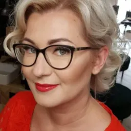 Portret kobiety w okularach, z profesjonalnym makijażem i upiętymi blond włosami, ubrana w czerwoną koronkową bluzkę.