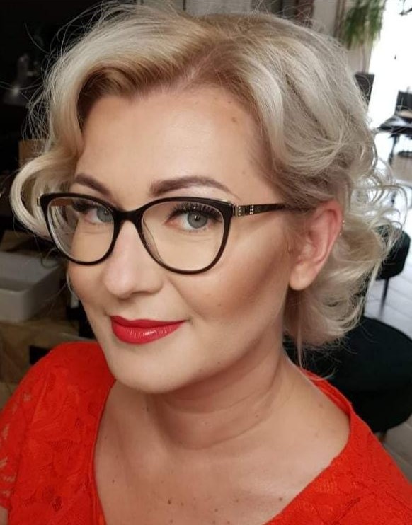 Portret kobiety w okularach, z profesjonalnym makijażem i upiętymi blond włosami, ubrana w czerwoną koronkową bluzkę.
