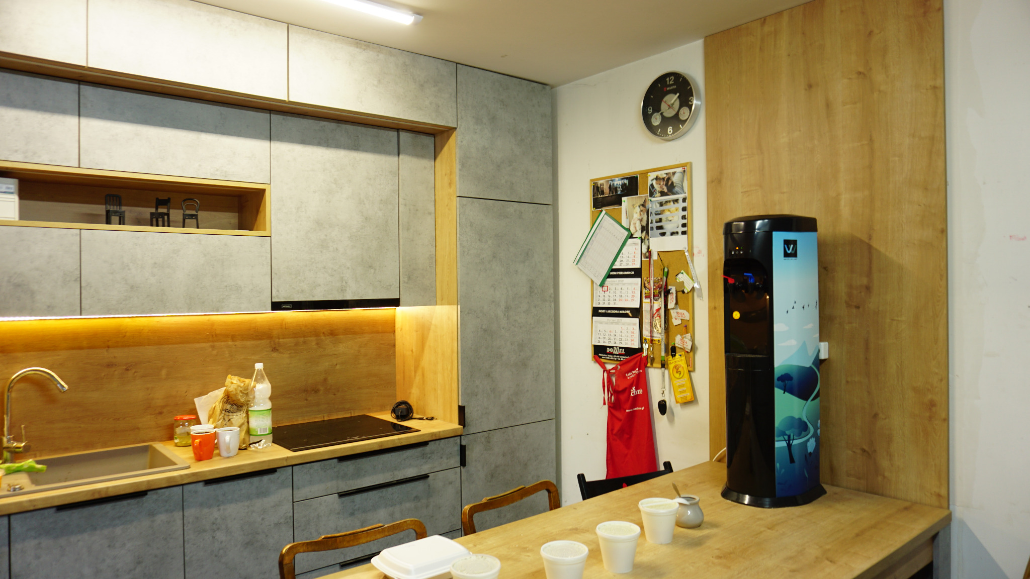 Wnętrze biura z szarymi szafkami kuchennymi, drewnianym blatem, dystrybutorem wody z grafiką górską i tablicą korkową z kalendarzami.