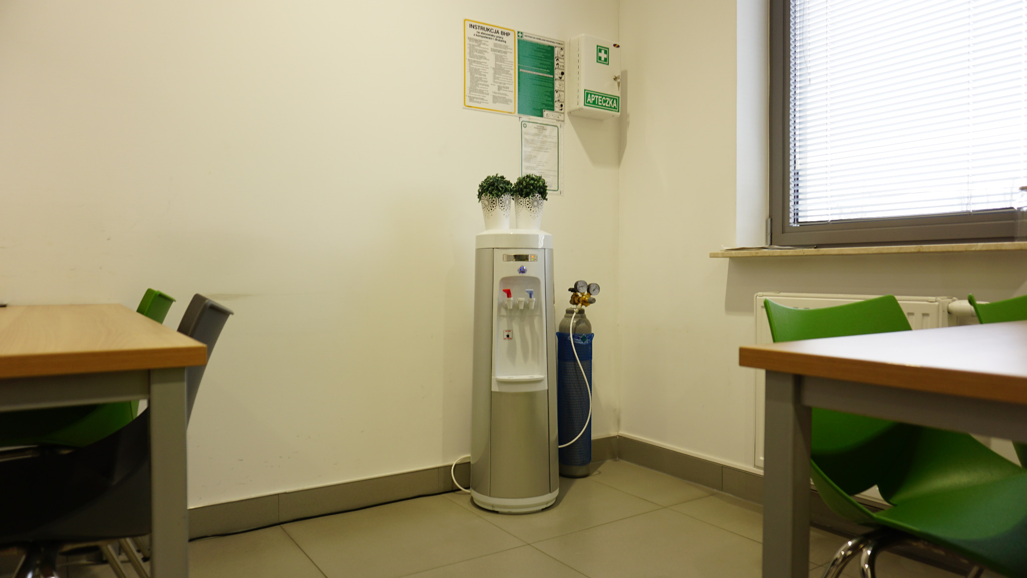 Srebrny dystrybutor wody z butlą gazową obok, ustawiony w rogu pomieszczenia biurowego z instrukcją BHP i apteczką na ścianie, oraz stołami z kolorowymi krzesłami.