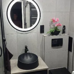 Nowoczesna łazienka w szarościach z czarnymi akcentami: okrągłe lustro z podświetleniem LED, umywalka nablatowa, wisząca toaleta, płytki imitujące beton i dekoracyjny akcent w postaci różowych...