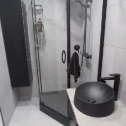 Nowoczesna łazienka z czarną kabiną prysznicową typu walk-in, czarną umywalką nablatową i szafką wiszącą, wykończona szarymi płytkami.