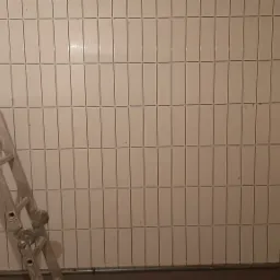 Ściana wyłożona wąskimi, pionowymi, białymi płytkami, z widoczną aluminiową drabiną i fragmentem płyty kartonowo-gipsowej w lewym dolnym rogu oraz podpórką teleskopową po prawej stronie, ukazująca...