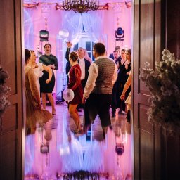 Bella Arte Show - Sala balowa z parkietem odbijającym światła, goście w strojach wieczorowych tańczą przy muzyce, widok zza dekoracyjnych kwiatów przy wejściu.