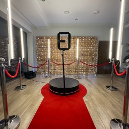 Bella Arte Show - Oświetlona platforma obrotowa 360 stopni z czerwonym dywanem i słupkami odgradzającymi na tle drewnianej ściany z lampkami.