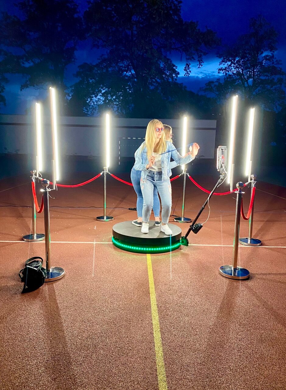 Dwie kobiety pozują na obrotowej platformie z oświetleniem LED i czerwonymi barierkami, robiąc sobie zdjęcia telefonem na statywie, na tle boiska.