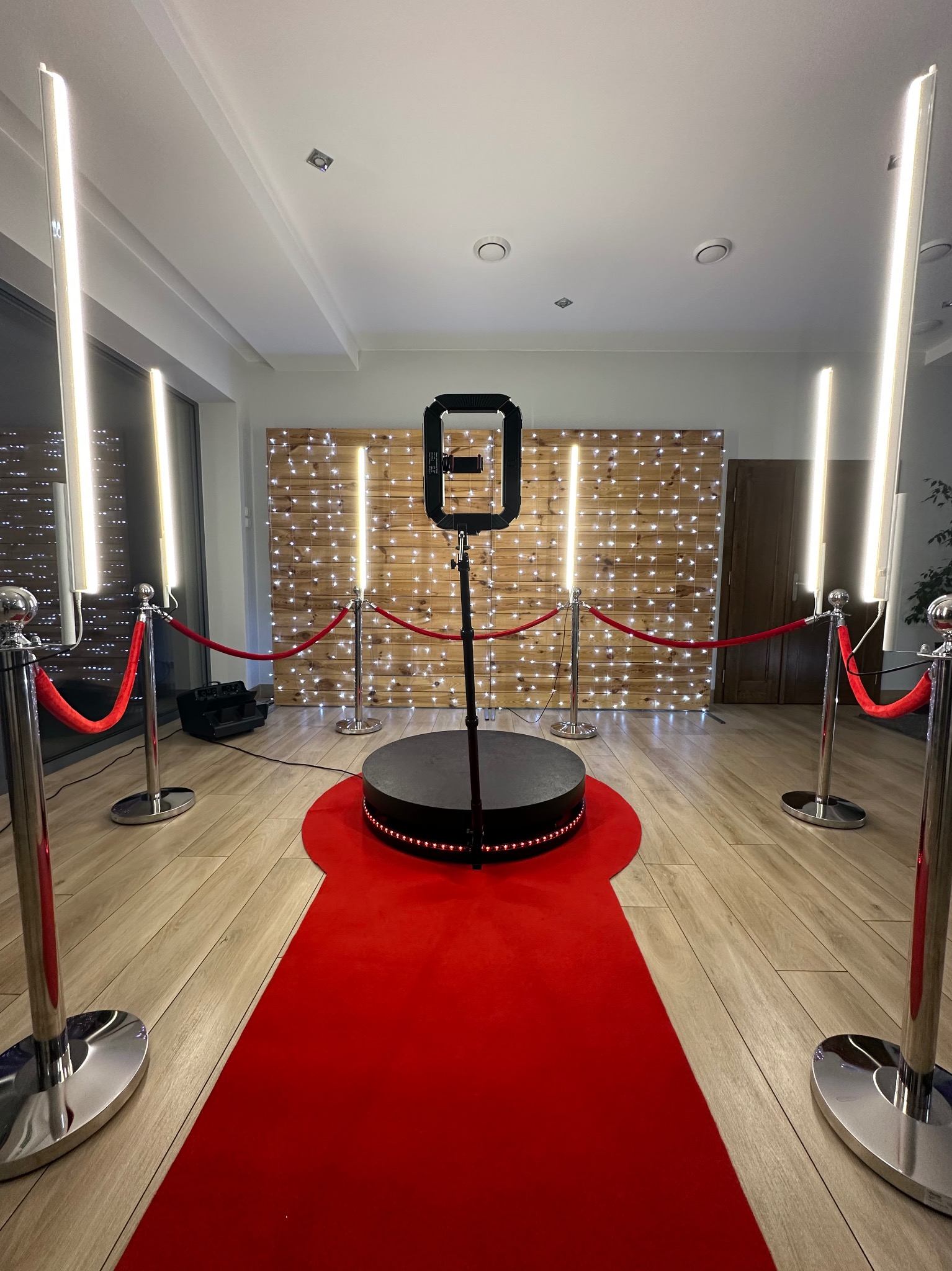 Oświetlona platforma obrotowa 360 stopni z czerwonym dywanem i słupkami odgradzającymi na tle drewnianej ściany z lampkami.