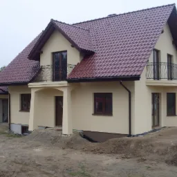 Beżowy dom jednorodzinny z ciemnoczerwoną dachówką i balkonami z kutymi balustradami, świeżo po pracach elewacyjnych, widoczne nierówności terenu wokół budynku.