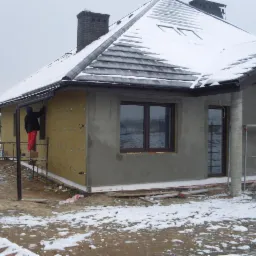 Ocieplanie elewacji domu jednorodzinnego w trakcie prac zimowych, widoczna wełna mineralna z kołkami mocującymi, częściowo otynkowana ściana, fragment rusztowania.