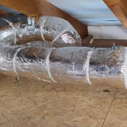 Izolowany kanał wentylacyjny z folii aluminiowej, przymocowany białymi opaskami zaciskowymi, biegnący pod drewnianym stropem na poddaszu.