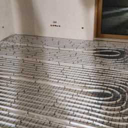 YOUVENT Michał Graczek - Białe rury ogrzewania podłogowego ułożone na czarnej siatce w jasnym pomieszczeniu z drewnianymi oknami. Widok z góry na instalację przed wylaniem posadzki.